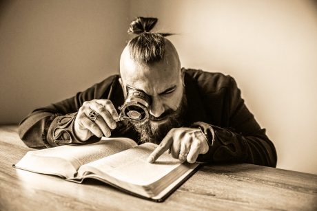 Ein bärtiger Mann betrachtet mit einer Lupe ein Buch. Er zeigt dabei auf einen Absatz.